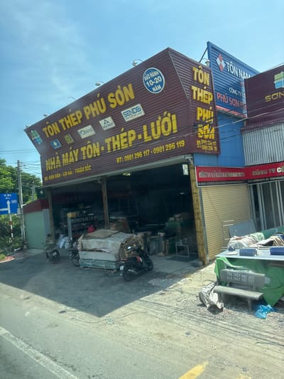 The image shows a storefront with prominent signage for "TÔN THÉP PHÚ SƠN," which appears to be a business dealing with roofing and steel products. The building has a multi-colored metal exterior, with a deep red section on the left and a bright blue section to the right. Large yellow lettering spells out "TÔN THÉP PHÚ SƠN" on the red facade, along with logos for brands like SENDO, POMINA, and HOA PHAT. A smaller sign below reads "NHÀ MÁY TÔN - THÉP - LƯỚI" (Steel Sheet - Steel - Mesh Factory). Further text on the building indicates phone numbers and addresses, including "ĐC: KIÊN LƯƠNG - LÔI HẢI - THUẬN BẮC".
The interior of the storefront is visible through an open entrance, revealing shelves stocked with various items and some equipment. Outside the shop, several motorcycles are parked, and materials like pallets, tarps, and what appear to be building supplies are stacked. In the foreground, there is a concrete bench with "TÔN THÉP PHÚ SƠN" written on it. A street sign with "TP. HỌC" and an arrow pointing to the right is visible on the left side of the image. The environment appears to be an outdoor commercial area with a paved road and some vegetation. The sky is visible, indicating it is daytime with clear weather.