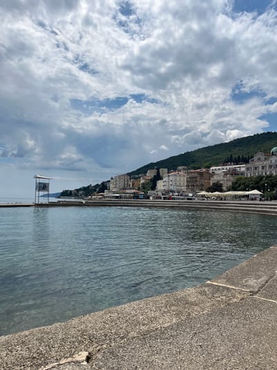 The image shows a view in the city of Grad Opatija, Croatia. The scene is primarily comprised of water, extending from the foreground towards a distant coastline featuring buildings and a verdant hillside. The right side of the frame contains a textured, concrete or stone barrier. The sky is filled with numerous clouds. A small structure or platform is situated in the water near the shoreline.