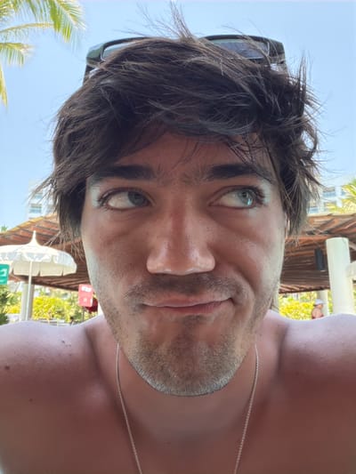 The photograph presents a close-up, tilted view of a man with fair skin, dark hair, and a light beard. He has light-colored eyes. A pair of sunglasses is perched atop his head. He wears a thin silver chain around his neck. The background reveals a bright, outdoor setting with palm trees and building structures, suggesting a tropical or resort location. The environment, including sun umbrellas and greenery, hints at a tourist area. The location is Las Jarretaderas, Mexico.
