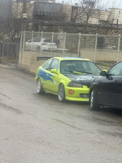 The scene depicts a street in Mas'ade, Israel, under overcast conditions. The foreground consists of a dark gray, damp paved surface. Two vehicles are parked closely together. On the left is a highly customized, two-door, lime green vehicle, resembling a Honda Civic. It features a black hood with white stylized lettering, blue lightning bolt graphics extending from the side into the rear quarter panel, a black rear spoiler, a sunroof, and silver multi-spoke aftermarket wheels. A red "HELLA" sticker is visible on the front bumper. To its immediate right, the front passenger side of a black compact vehicle, possibly a hatchback, is parked, showing a silver-spoked wheel. In the midground, a gray metal mesh fence with white support posts runs along the top of a concrete retaining wall. Behind this fence, a white SUV is partially visible. The background includes leafless trees and several buildings with light-colored stone facades. A utility pole with a small white component is present on the far left. No people are visible.