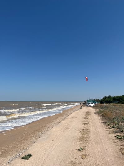 The image depicts a view of the Sea of Azov coastline near Dolzhanskaya, Russia. A sandy beach borders the choppy, brown water. A dirt road runs parallel to the beach, with dry grass and sparse vegetation on its right side. A white car is parked on the side of the road near a structure covered with a green tarp. A bright red kite is visible in the clear blue sky. The scene suggests a daytime environment with strong sunlight, typical of a summer day.