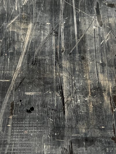 The image depicts a close-up, textured view of a heavily scratched and worn dark surface, possibly wood or laminate, exhibiting vertical grain patterns and numerous parallel gouges and scuffs. Streaks of lighter material, likely dirt or residue, are embedded within the scratches, creating a distressed appearance. Several dark, irregular splotches are also visible on the surface, some appearing to be areas of deeper damage or accumulated grime. The overall impression is one of age and heavy use, without any identifiable context or specific objects other than the textured surface itself. The image does not provide any information about the city of Lanžhot, Czechia, nor does it contain any discernible human presence or activity.