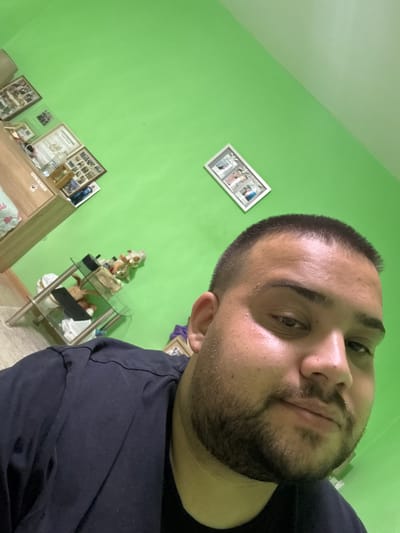 A man is taking a selfie in a room. The room has bright green walls and a wooden shelf unit with framed pictures. A glass-top side table with stuffed animals is also visible. The man is wearing a dark t-shirt and has short hair and a beard. The image is taken from a close-up perspective, focusing on the man's face and upper body. The location is specified as Byala Reka, Bulgaria.