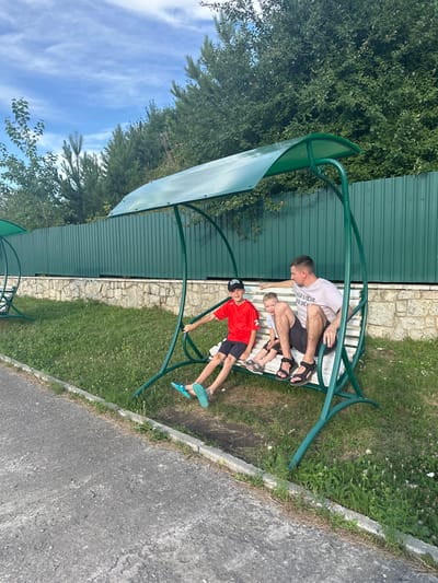 A father and his two sons are sitting on a green metal swing set with a green canopy. The father is wearing a grey t-shirt and black shorts, and the older son is wearing a red t-shirt and black shorts with a black baseball cap. The younger son is wearing a light blue t-shirt and grey shorts. The swing set is on a grassy area with a stone wall and a green metal fence behind it. Trees are visible in the background. The scene appears to be outdoors, likely in a park or recreational area, in Даниловка (алтайский), Russia. The ground in front of the swing set is paved with asphalt.