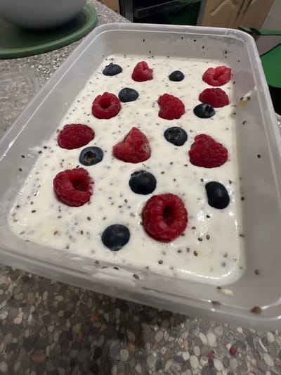 A rectangular, transparent plastic container filled with a creamy white substance, possibly yogurt or chia seed pudding, is shown in the image. The pudding is speckled with small, dark seeds. Adorning the top of the mixture are several fresh raspberries and blueberries, arranged seemingly at random. The container appears to be resting on a countertop with a speckled grey and white pattern. A green bowl and lid are visible in the lower-left corner of the frame. The image does not contain any human subjects. The location is not specified in the image, and thus cannot be pinpointed to Blackburn, United Kingdom.