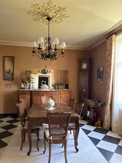 This image captures a traditional and elegantly furnished dining room, likely within a home in Étréchy, France. The room is characterized by warm, light tan walls with white decorative molding and a rich, dark wooden aesthetic for its furniture.

In the center, a large, ornate dark wood dining table is surrounded by matching chairs featuring cane backs and light, padded seat cushions. Above the table hangs a prominent black wrought-iron chandelier with white lamp shades, suspended from an intricate white ceiling medallion.

Along one wall, a tall wooden grandfather clock with a white face and dark Roman numerals stands next to a bar cart laden with various bottles and glasses. Opposite this, a large, ornate wooden sideboard or cabinet with carved details is positioned beneath a multi-paneled mirror, which reflects the chandelier and parts of the room. A vase with flowers and greenery is displayed on the sideboard.

On the walls, several framed pictures are visible: one above the grandfather clock and two smaller ones lower down, including one depicting an animal resembling a deer. An decorative white urn or tureen serves as a centerpiece on the dining table.

The flooring consists of a striking black and white checkerboard tile pattern in some areas, transitioning to a lighter-colored rug or carpet under the dining set. A window with white panes and a patterned valance can be seen at the top of the image.

No specific activity is taking place; the room appears set up for daily use or a gathering. A person is partially visible in the reflection of the large mirror, appearing to wear a dark top, but their features are indistinct. The lighting suggests daytime. There is no discernible text visible in the image.