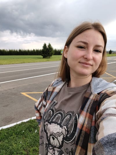 A woman is taking a selfie outdoors. She is wearing a brown t-shirt with a graphic print, and a plaid flannel shirt is draped over it. The background features a paved road with white and yellow markings, a green field, a row of coniferous trees, and a cloudy sky. A road sign is also visible in the background. The location is Горняк, Russia.