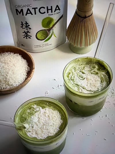 The image presents a top-down view of a still-life arrangement featuring ingredients and prepared drinks related to matcha. Two clear glasses, positioned in the upper right and lower right, contain layered green matcha lattes, generously topped with white shredded coconut flakes. Each glass has a clear glass straw inserted.

To the left, a traditional bamboo matcha whisk (chasen) rests on a light green ceramic whisk holder. Below the whisk, a white pouch of matcha powder is visible. The pouch is prominently labeled "ORGANIC MATCHA" in large black letters and states "Made in Japan." Further details on the pouch include "VEGAN," "GLUTEN FREE," "ORGANIC," "NON GMO," and "80g," along with Japanese characters "抹茶".

A small, natural wooden bowl filled with shredded coconut flakes is situated below the matcha pouch, to the bottom left of the glasses. Sparse white coconut flakes are scattered across the clean, white surface that forms the background for the entire composition. The scene suggests a curated display or preparation of coconut matcha lattes, photographed indoors under soft, even lighting. No people are visible, and there are no discernible location cues beyond the product's origin.