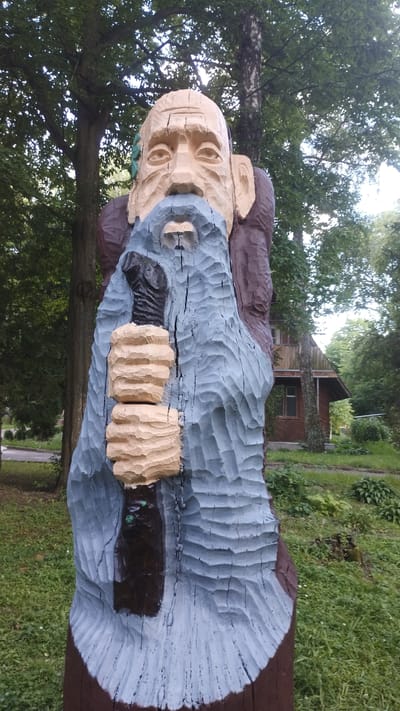 A wooden sculpture of an old man with a beard is standing in a park. The sculpture is roughly carved and painted with blue, brown, and beige colors. The man has a weathered face with deep-set eyes and a long, flowing beard. He is holding a staff in his hands. The sculpture is set against a backdrop of trees and a small wooden house in the distance. This sculpture is located in the Park of Culture and Leisure in Khmilnyk, Ukraine.