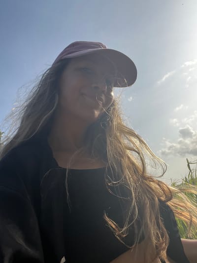 A low-angle selfie shows a young woman wearing a pink baseball cap and a black top. She has long, light brown hair that is being blown by the wind. The background is a bright blue sky with scattered white clouds. The sun is shining, creating a lens flare on the right side of the image. The woman is smiling and looking slightly off-camera. The overall impression is one of a casual, outdoor portrait. The location is Talalla, Sri Lanka.