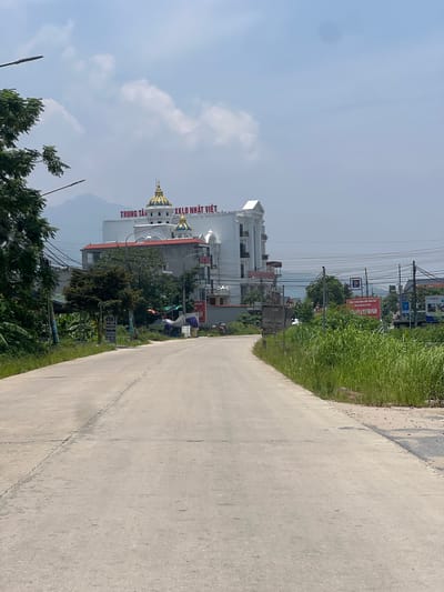 In Tu Vũ, Vietnam, a paved road curves to the right, flanked by lush green vegetation on the right and trees and buildings on the left. In the distance, a large white building with distinctive golden domes stands out against a bright, cloudy sky. Red lettering on the building reads "TRUNG TÂM XKLD NHẬT VIỆT". Further to the right, a red sign advertising "SHOWROOM" is visible amidst the greenery. Electrical wires crisscross the sky, and a few parked motorcycles suggest a normal day in this roadside scene. The overall impression is one of a developing area with commercial activity.