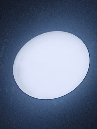 An oval, white egg rests on a dark blue, textured surface. The egg is centrally placed and illuminated, casting a soft glow around its edges. The background is uniformly dark blue and slightly out of focus, with a subtle grainy texture. There are no people or other objects visible in the scene. The image is a close-up, top-down view of the egg. The specific location in the city of Aygezard, Armenia, is not discernible from the visual information provided.