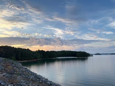 A wide, tranquil lake is shown at dusk, with a tree-lined shore and a rocky embankment in the foreground. The sky is a mix of blue and white clouds, with the last rays of sunlight casting a warm glow. The scene is serene and peaceful, with no visible signs of human activity other than a few distant figures on the shoreline. The location is likely Lake Lanier in Sugar Hill, Georgia, a popular recreational area known for its natural beauty and outdoor activities.
