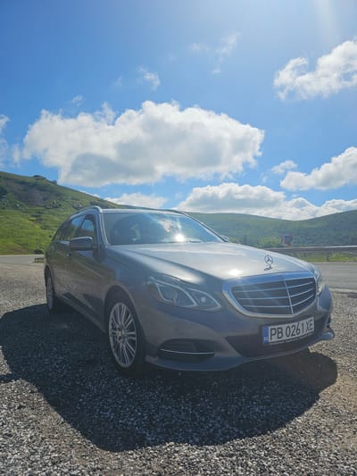 A grey Mercedes-Benz E-Class station wagon is parked on a gravel shoulder next to a road, with a grassy, hilly landscape and a bright blue sky with white clouds in the background. The car is facing slightly to the left, with its front grille and headlights clearly visible. The license plate reads "PB 0261 XE" and shows "BG" for Bulgaria. The surrounding environment appears to be a rural or mountainous area, with the hills covered in green vegetation. There is a guardrail along the edge of the road. The lighting suggests it is daytime, likely with the sun high in the sky. The image does not contain any people or specific landmarks that would definitively place it in Hristo Danovo, Bulgaria, though it is consistent with a rural Bulgarian landscape.