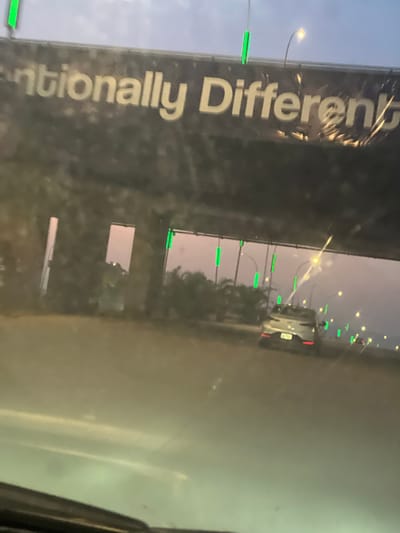 The image depicts a roadside scene at dusk or night in Kunkwaba, Nigeria. On the left, a large, dark banner or billboard is prominently displayed vertically. White capital letters are visible on the banner, spelling out the phrase "Intentionally Different". The surface of the banner appears somewhat reflective and crinkled in places.
To the right of the banner, a road stretches into the distance, lined with numerous streetlights emitting a warm, yellowish-orange glow. Distinctive bright green LED lights are mounted on poles along the roadside, appearing as vertical segments that punctuate the view. A white car, possibly an SUV, is visible further down the road, its form illuminated by the surrounding lights. The sky above is a pale grey-blue, indicating either late evening or early morning. The overall setting suggests an urban or semi-urban environment, with artificial light sources dominating the visibility. No people are discernible in the frame.