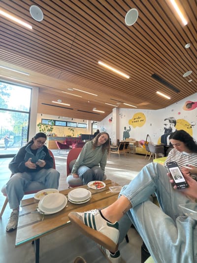 A candid, wide-angle shot captures three young people gathered around a table in a modern, well-lit common area in Kiryat Malakhi, Israel. The image is tilted at a Dutch angle.
The setting features a striking wooden slatted wall and ceiling on the left, integrated with linear strip lights and circular recessed lights. On the right, a white wall is adorned with colorful, stylized murals, including abstract shapes, figures, and a prominent black and white portrait-like depiction of a person. Within a yellow speech bubble on the wall, Hebrew text is visible, reading "אנשים הזזים את" (Anashim Ha-Zazim et), which translates to "People who move the..."
Three individuals are seated at a wooden table with a glass top. In the foreground, a person wearing glasses and a dark jacket holds an object in their hands. Across from them, another person with curly hair, wearing a striped long-sleeve shirt and light pants, smiles faintly towards the camera. On the far side of the table, a third person, partially visible, wears a striped shirt and light blue jeans, with white and green Adidas sneakers. They are looking at a smartphone, which displays a black and white image of a person.
Several white paper plates are on the table, some containing food, possibly salad. Large windows along the back wall let in bright natural light, indicating it is daytime, and offer a glimpse of green trees outside. The space appears to be a casual lounge, cafeteria, or community center, designed for social interaction.