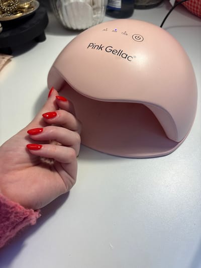 A hand with freshly painted red almond-shaped nails rests near a pink "Pink Gellac" UV nail lamp. The hand is positioned as if it is about to be placed under the lamp. A gold ring adorns the ring finger. The hand is casting a shadow on the white surface to the right of the lamp. Other beauty-related items are visible on the left, slightly out of focus. The sleeve of a pink sweater is visible at the bottom right corner.