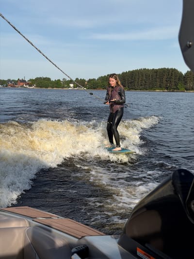 A woman is wakeboarding on a body of water in Терехово, Russia. She is wearing a wetsuit and is being pulled by a rope attached to a boat. The boat is visible in the right of the frame, and trees line the shore in the background. The sky is a pale blue. The woman is standing on the wakeboard with her feet bare.