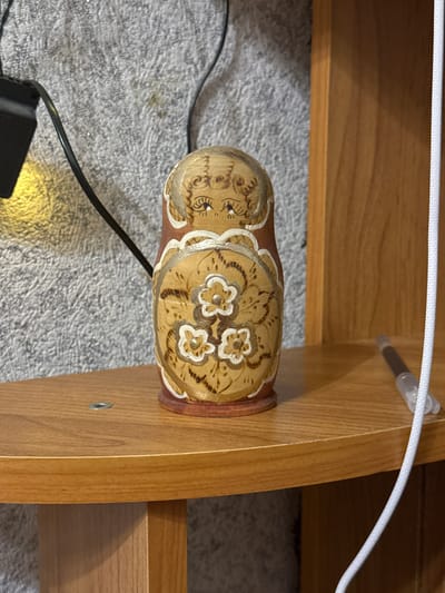 A single Matryoshka doll, painted with an intricate floral design and a stylized face, rests on a wooden shelf. The doll is brown and tan, with white accents on the flowers and face. To the left of the doll, a black electrical cord is visible, attached to a black power adapter that emits a soft yellow light. The background is a textured, gray wall, possibly a carpet or fabric. To the right of the doll, a white cord runs vertically, and a pen with a clear plastic tip is lying on the shelf. The shelf itself is made of polished light brown wood, with a visible metal screw in the center. The overall scene appears to be indoors, likely a domestic setting. This scene is located in Масаловка, Russia.