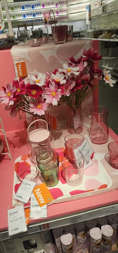 The image shows a retail display of glassware and artificial flowers in a store, likely an IKEA, in Gwangmyeong, South Korea. In the foreground, a pink tray with an abstract pattern holds several glass items, including green and clear tumblers and a small pink, flower-shaped dish. A pink lantern-like glass container with a handle is also visible. Behind the tray, a bouquet of pink and white artificial cosmos flowers is displayed in a clear glass vase, with another pink vase to its right. Several price tags and "New" tags are attached to the items, with some indicating prices in Korean Won. In the background, shelves are filled with various glassware and other retail items. The overall scene is brightly lit and organized, typical of a retail environment.