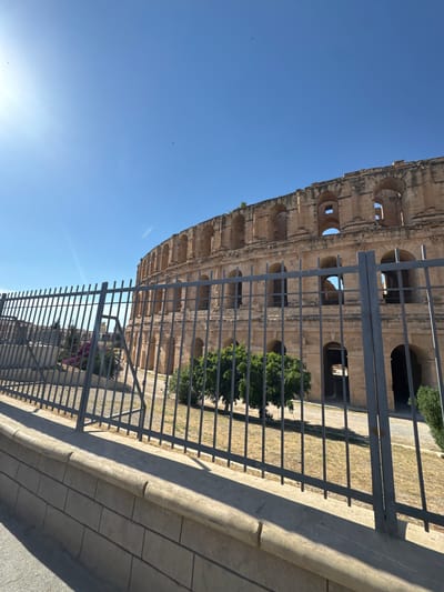 The image captures a partial view of the El Jem amphitheater in El Jem, Tunisia, from behind a gray metal fence. The amphitheater's facade is constructed from tan-colored stone with arched openings. A low wall with a stone ledge runs along the bottom edge. The sky is a clear, light blue. Vegetation, including green bushes, can be seen within the amphitheater's structure. The scene is bathed in daylight, casting shadows on the wall and ground.
