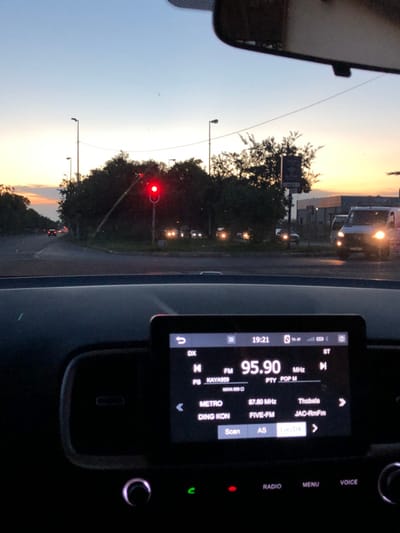 Inside a car, the dashboard displays a radio tuned to 95.90 MHz, identifying as KAYA 959, playing POP music. Alternate stations listed are Thobela, JAC-RmFm, FIVE-FM, and METRO. Outside the car, a traffic light is red, and streetlights illuminate a dusky scene. Trees line the road, and other vehicles are visible. A portion of the car's rearview mirror is also visible.