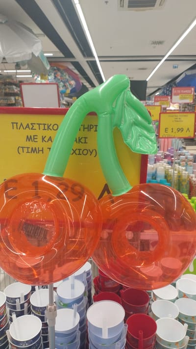 In Pylaia Municipal Unit, Greece, a brightly lit supermarket aisle features a display of inflatable cherry-shaped pool floats. The floats, primarily orange with green stems, are positioned prominently in the foreground. Behind them, a yellow sign with Greek text is visible. To the right of the floats, a price tag displays "E 1,99 A". The background reveals shelves stocked with various items, including stacks of colorful cups, suggesting a retail environment. The overall scene depicts a commercial setting focused on seasonal or novelty items.