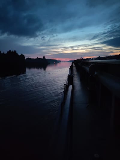 The image captures a twilight scene from the perspective of a vessel navigating a calm body of water, likely a river or canal, near Badozhskoi Pogost, Russia. The sky is a blend of deep blues and grays, with hints of orange and pink near the horizon, suggesting either dawn or dusk. Dark silhouettes of trees line the distant shore on the left. On the right side of the image, the dark, angular structure of the vessel itself occupies a significant portion of the frame, with railings and a walkway visible. The water shows gentle ripples. There are no people visible in the image.