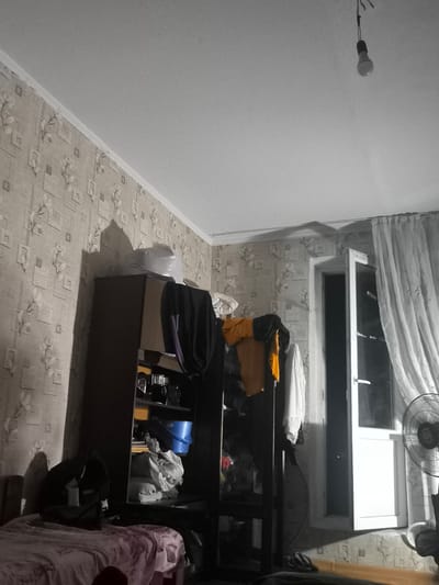 The image depicts a dimly lit room, possibly a bedroom or living space, in Dubossary, Moldova. The setting appears somewhat cluttered, with a large, dark wardrobe or shelving unit dominating the left side of the frame. Various items, including clothing, bags, and what looks like a blue bucket, are visible on and around the shelves.
A bed covered with a pinkish cloth is partially visible in the foreground on the left. The walls are covered with patterned wallpaper. An open window or door on the right side of the image reveals a white door and a curtain. The light from outside is bright, casting a shadow of the window frame and curtain onto the wall. A standing fan is positioned to the right of the window.
A single light bulb hangs from the ceiling by wires, providing the primary illumination in the room, suggesting it is nighttime or the room is otherwise dark. The overall mood is somewhat stark and utilitarian, with no clear indication of a specific activity or event taking place. The time of day is likely evening or night, given the artificial lighting and the contrast between the interior and exterior light. There is no visible text in the image.