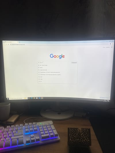 The image shows a computer monitor displaying the Google search page. The browser tab is in Russian and reads "New tab". Below the search bar, there are several suggested searches, also in Russian, including "Suzuki Jimny 1.3 AT reviews for engine and gearbox". The monitor is positioned on a desk, with a white keyboard with RGB backlighting in the foreground. To the left of the monitor is a white mug, and to the right, a stand for the monitor is visible. The overall environment appears to be an indoor setting, possibly a home office or study. The date and time visible in the lower right corner of the screen are 15:41, 07.07.2026. There is no information in the image to suggest a specific location within Saraktash, Russia.