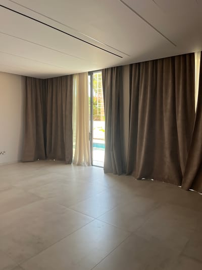 The image depicts an interior view of a room with large sliding glass doors leading to an outdoor pool area. The room has light-colored tiled flooring and cream-colored walls. Two sets of curtains are visible: dark brown, heavy velvet curtains on the right and in the left foreground, and lighter, sheer curtains partially visible on the left. The ceiling is white with subtle linear detailing and integrated lighting. Through the glass doors, a swimming pool with blue water and some surrounding greenery are discernible. The scene is clean and appears to be a residential property in Moni Community, Cyprus, likely a living area or bedroom with a view of the pool. There are no people or animals present in the image.