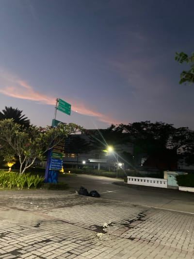 The image depicts a street scene at dusk in Mekar Bakti, Indonesia. The sky displays a gradient of deep blue to purple with streaks of pink clouds. A directional signpost with green signs indicating "Curug" and "Cikupa" is prominently visible on the left side of the road, along with a blue sign listing various locations such as "OFFICE PARK," "ECO PLAZA," and "SEKOLAH TARAKANITA." In the foreground, a paved area with brick-like patterns shows damp patches and scattered debris. Two black trash bags lie on the paved surface. A streetlamp illuminates the area, casting a bright glare. Lush green plants and a tree with pink flowers are situated to the left, while a large, dark silhouette of a tree stands on the right. In the background, a building with a prominent "CGV" logo is visible, suggesting it's a cinema or entertainment complex, with other structures and trees forming the urban landscape. The scene is devoid of people.