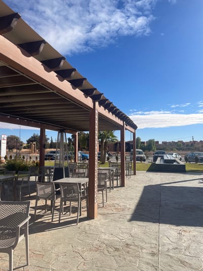 This image depicts an outdoor patio of a commercial establishment in Lkheng, Morocco, under bright daylight. A brown wood-composite pergola, featuring a light fabric canopy and decorative lattice-work side panels, covers an area with multiple unoccupied grey plastic tables and chairs arranged in rows. A tall, silver outdoor patio heater stands among the seating. The ground is paved with irregular grey and reddish-brown stones, displaying distinct shadows from the pergola structure and furniture. Beyond the patio, a street is visible, separated by a landscaped median containing green shrubs and palm trees. Several vehicles, including cars, vans, and a motorcycle, are present on or near the street. A dark, rectangular water feature with multiple spouts is situated to the right of the street. Distant low-rise buildings and additional vegetation are visible in the background under a clear blue sky with scattered white clouds. A partial white "Toyota" sign is visible on the far left. No individuals are present within the immediate patio area.