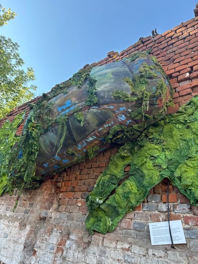 An outdoor art installation is displayed on a brick wall in the city of Yelets, Russia. The installation features a large, cylindrical object, possibly resembling a pipe or a tunnel, covered in green fabric that mimics moss or foliage. This green material hangs down in tattered strips. The cylindrical object itself is painted with abstract patterns of blue, brown, and dark green, suggesting a weathered or naturalistic texture. The brick wall supporting the installation is weathered and shows signs of age, with some sections appearing damaged or crumbling. A small white sign with text in Russian is attached to the wall below the artwork. In the upper left corner of the image, a portion of a green leafy tree is visible against a clear blue sky. Two small, dark, bird-like figures are perched on the top edge of the brick wall.