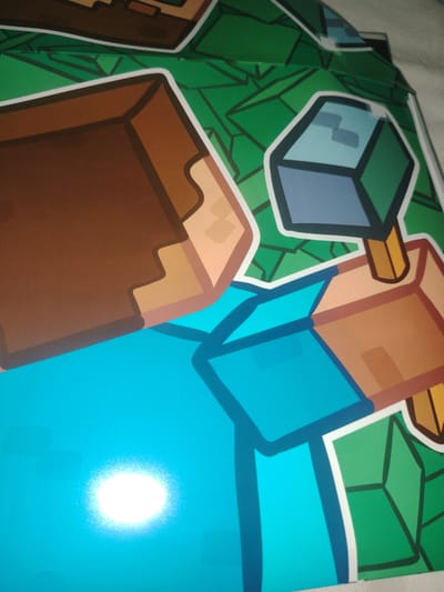 The image shows a close-up of a Minecraft-themed graphic. The graphic features blocky, pixelated characters and objects typical of the game, rendered in bold outlines and flat colors. Dominant colors include brown for blocky textures, bright blue for character clothing or body parts, and various shades of green for the background, which appears to be a stylized representation of grass or foliage with geometric patterns. The lighting in the image suggests it might be indoors, with a bright, possibly artificial, light source causing a glare on the blue surface. The image is a partial view, showing portions of two figures, one with a brown blocky arm and the other holding a blue and grey cube object. The overall impression is of a colorful and stylized depiction of elements from the video game Minecraft. The image is not situated in Xonobod City, Uzbekistan; it is a generic graphic representation.
