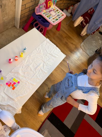 A young girl is indoors, standing over a white table. On the table is a length of white material, likely fabric, with colorful painted squares at one end and containers of paint nearby. The girl is holding a paintbrush and has one hand in her overall pocket. The surrounding environment includes a toy learning center, wooden flooring, and a rug. The location of Bearpark, United Kingdom, is not discernible in the image.