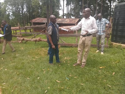 The scene depicts four African men in a grassy area. One man on the left is walking away while talking on a cell phone and holding a document. Two men are conversing near a rustic wooden fence and shed, one gesturing with a paper in his hand. A fourth man stands with his hands on his hips near a large, black water tank. The location appears to be an outdoor setting, possibly a farm or rural area, given the presence of the wooden fence, shed, and tall trees in the background. The presence of the water tank suggests a focus on water management in the area. The image does not contain any specific details to confirm the exact location within Idakho South ward, Kenya.