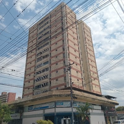 The image displays a daytime, low-angle view of a multi-story building identified as Edificio Don Bosco, located at Avenida Los Leones with Carrera 18, Barquisimeto, Lara State, Venezuela. The prominent structure is a tall, approximately 20-story building featuring a light beige facade with distinct horizontal reddish-brown bands on each floor. Numerous rectangular windows are visible, some with external air conditioning units. The building's lower two levels house commercial establishments, characterized by a lighter grey and blue segmented facade and an extended concrete overhang. Multiple "SKF" signs are affixed to the commercial section, one explicitly stating "Rodamientos Transmisión Industrial" and another displaying a partial phone number "4412404." Overlapping the foreground and midground are numerous black overhead electrical or communication cables, strung between utility poles, partially obscuring the building and the partially cloudy blue sky. Green foliage from trees is visible on the left and right sides of the lower frame. A shorter, reddish-pink building is visible in the background to the far left. Two vehicles, one white and one dark, are partially visible on the far right. No discernible human figures or specific interactions are present within the frame.