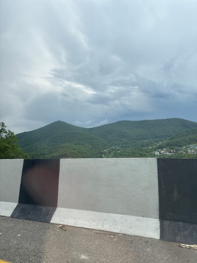 The image depicts a roadside view with a concrete barrier in the foreground and lush green mountains in the background. The sky is overcast with heavy clouds. The barrier is painted with alternating black and white stripes. Scattered houses and buildings are visible nestled on the slopes of the mountains. The scene appears to be a natural landscape with some human habitation, possibly a rural or suburban area. The location is identified as Dederkoi, Russia. The image composition suggests the photo was taken from a moving vehicle, as indicated by the angle and the slight blur on the roadside. There are no people or animals visible in the frame.