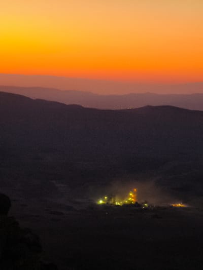 This image captures a remote scene at dusk, likely in or around the city of Yeruham, Israel. The sky transitions from a bright, warm orange at the top to softer hues of pink and then a deep purple as it approaches the horizon. Layers of distant, dark mountains are silhouetted against the sky, progressively fading into the darkness.

In the foreground, a cluster of lights illuminates a settlement or industrial area. The lights are a mix of yellow and orange, suggesting artificial illumination. There is a haze or mist surrounding these lights, adding to the atmospheric and somewhat mysterious quality of the scene. The terrain immediately surrounding the lights appears to be dark and rugged, consistent with a desert or semi-arid environment. On the far left of the image, a dark, silhouetted object, possibly a rock formation or a person, is visible in the foreground. The overall impression is one of isolation and the presence of human activity in a vast, natural landscape.