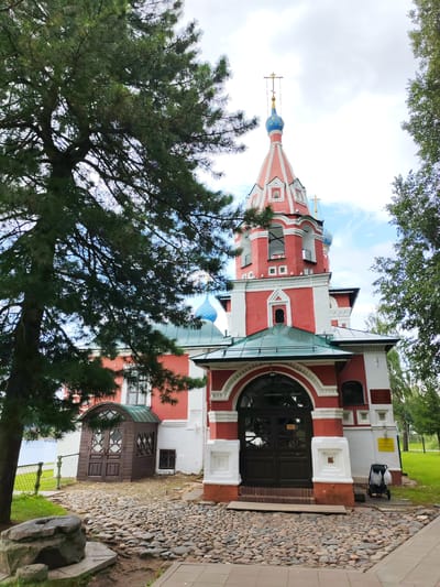 The image shows the Church of the Assumption of the Blessed Virgin Mary in the city of Vasil'ki, Russia. The church is painted red and white, with a distinctive bell tower topped with a blue dome and a golden cross. The entrance to the church is through a large arched doorway with dark wooden doors. In front of the church is a cobblestone courtyard. To the left of the church is a small wooden structure with diamond-shaped windows. A large pine tree is visible in the foreground on the left side of the image, its branches partially obscuring the church. The sky is cloudy, with patches of blue visible. There are no people visible in the image.