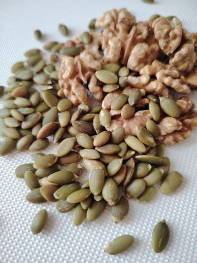 This media file is a close-up, eye-level shot of a pile of shelled pumpkin seeds and walnuts on a white, textured surface. The pumpkin seeds are predominantly a muted green color, with some appearing lighter and more translucent, while others are a darker green or brown. They are scattered and overlapping, creating a textured foreground. Behind the seeds, a cluster of walnuts is visible. The walnuts are a light brown color, with their characteristic convoluted surface clearly defined. The lighting is bright and even, suggesting an indoor setting with good illumination. There are no people or discernible activity other than the static arrangement of the food items. The white surface has a subtle, regular texture, possibly a cloth or paper. The overall impression is one of a simple, natural composition focusing on the textures and colors of the nuts and seeds. No text is visible in the image.