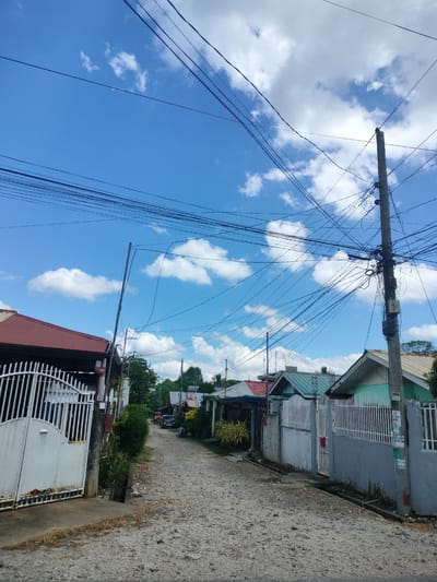 The image depicts a residential street in Kidapawan City, Philippines, on a bright, sunny day with scattered clouds. A gravel-covered road stretches into the distance, lined with houses and fences. On the left, a large white metal gate guards the entrance to a property with a red-roofed house. Various electrical wires are strung across the sky, supported by concrete utility poles. The houses visible have a mix of metal and tiled roofs in red, green, and blue. Green foliage and trees are interspersed among the structures. A few vehicles, including a car and a motorcycle, are parked along the street. The scene conveys a typical urban or suburban environment in the Philippines.