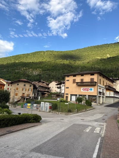 The image depicts a street scene in Spormaggiore, Italy. The perspective is from a slightly elevated position, looking down a winding road. On the left side of the road, a steep, heavily forested mountainside dominates the scene, with a bright blue sky dotted with white clouds above it. A faint contrail from an airplane is visible in the sky.
The right side of the road features a cluster of buildings. The most prominent building is a multi-story structure with a light orange facade, featuring several windows and balconies. A large, red and white "coop Trentino" logo is prominently displayed on this building, indicating a retail cooperative. Below this building, other structures are visible, suggesting a small town or village. The road itself is paved, with white pedestrian crossing markings and various signs. To the far right, a brick-paved sidewalk runs alongside the road.
In the foreground, there is a green grassy area with some bushes and trees. There are also several bins for waste separation visible near the buildings. The overall impression is of a quiet, picturesque mountain village on a clear day. The time of day appears to be daytime, likely late morning or early afternoon, given the bright sunlight and shadows. There are no people visible in the image.