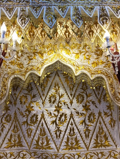 The image shows a close-up of an ornate, white fabric with extensive gold embroidery. The embroidery features floral and leaf motifs arranged in a repeating pattern. Decorative gold bead fringe hangs from scalloped edges in the upper portion of the fabric. Two crystal sconces with lit bulbs are visible on either side of the frame, attached to what appears to be a curtain rod or decorative fixture. The overall impression is one of richness and intricate craftsmanship. The location, Cottrueng, Indonesia, is noted as context but is not visually represented in the image itself.