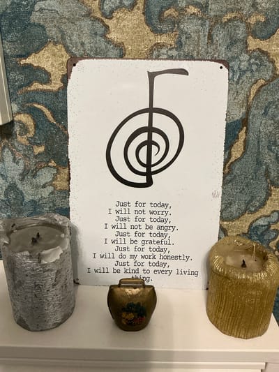This media file displays a white metal sign with black text and a graphic symbol, placed on a patterned rug. Two candles, one gold and one silver, are positioned on either side of the sign, and a small decorative object with floral imagery sits atop the gold candle. The rug features a design with shades of blue, tan, and gold, creating a textured background.

The text on the sign reads: "Just for today, I will not worry. Just for today, I will not be angry. Just for today, I will be grateful. Just for today, I will do my work honestly. Just for today, I will be kind to every living thing." The graphic symbol on the left side of the sign appears to be a stylized representation of the "Enso" circle, a Buddhist symbol.

The scene is indoors, likely on a flat surface like a table or shelf, given the presence of the rug and the arrangement of the objects. The lighting is soft, suggesting it could be daytime with natural light, or artificial lighting. There are no people visible in the image. The overall mood is one of tranquility and mindfulness, suggested by the text on the sign and the presence of candles, which often symbolize peace and reflection. The location context of Bielefeld, Germany, does not provide any specific visual cues within the image itself.