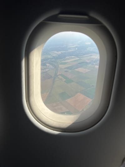 An airplane window frames a patchwork of agricultural fields below, seen from above. The fields are predominantly green and brown, suggesting cultivated land. A road or river winds through the landscape. The view is slightly hazy, possibly due to altitude or atmospheric conditions. Sunlight streams onto the window frame, creating bright highlights and deep shadows, adding depth to the composition.