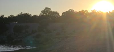 A wide shot shows a bright sun setting over a treeline on a sandy hill. The sun is a large, glowing orb in the upper right corner of the frame, with rays of light fanning out and creating a lens flare effect. Below the treeline, the sand appears to be a light tan color, with some darker patches of vegetation. In the foreground, a body of water, likely a lake or river, reflects the light of the setting sun, creating a shimmering surface. The overall impression is one of a serene natural landscape at dusk. The location is the city of Issyk-Kul, Kyrgyzstan.