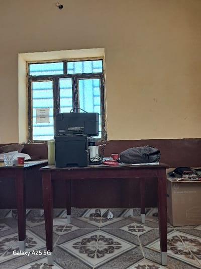 The image shows a room with a window, a desk with a computer on it, and a tile floor. The room is located in Karima, Sudan, at the coordinates 18.54 latitude and 31.831 longitude.  The desk is made of wood and is brown. The computer is black and has a monitor on top of it. The room has a window with a blue sky visible through it. The room has a tile floor. There are two chairs on the other side of the desk, obscured by the desk. There is a brown bag on the desk. There is a box on the floor next to the desk. The box is white with black lettering. There is a paper cup on the desk. The walls are beige. There are no people in the image.