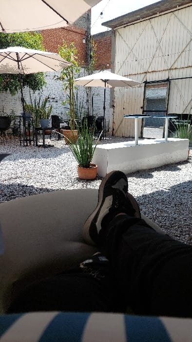The image captures an outdoor patio in Tlaxcala City, Mexico, from a low-angle perspective. The foreground displays the legs and feet of an individual, dressed in dark pants and black and white athletic shoes, resting on a surface patterned with blue and white stripes. The ground of the patio is covered with light gray gravel. In the midground, several dark metal tables and chairs are arranged alongside various potted plants, including tall, slender green foliage in terracotta pots. Two large, open light-colored patio umbrellas provide shade. A central, low, rectangular white concrete block structure, possibly a planter or bench, is visible, with two small tables featuring round, light-colored tops on white pedestals behind it. The background on the left consists of a white-painted brick wall partially obscured by greenery, with a section of exposed red brick higher up. On the right, a tall, white-painted building features a large, double-door structure with a prominent diagonal cross-hatch pattern and a small, dark-framed window. String lights are strung between the background structures. The scene is brightly illuminated by direct sunlight, casting distinct shadows. No other individuals are visible.